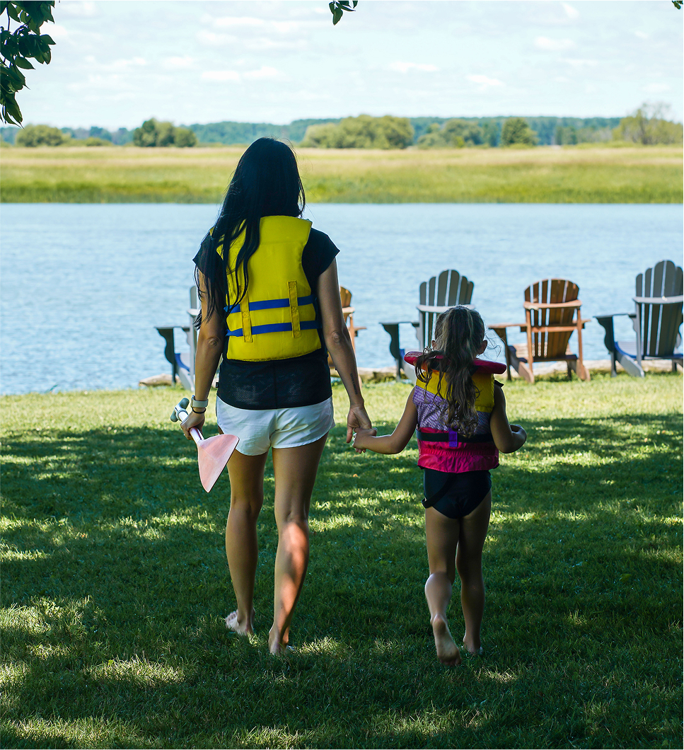 Mère et fille portant des gilets de sauvetage marchent main dans la main vers un lac bordé de chaises Adirondack, par une journée d'été ensoleillée.